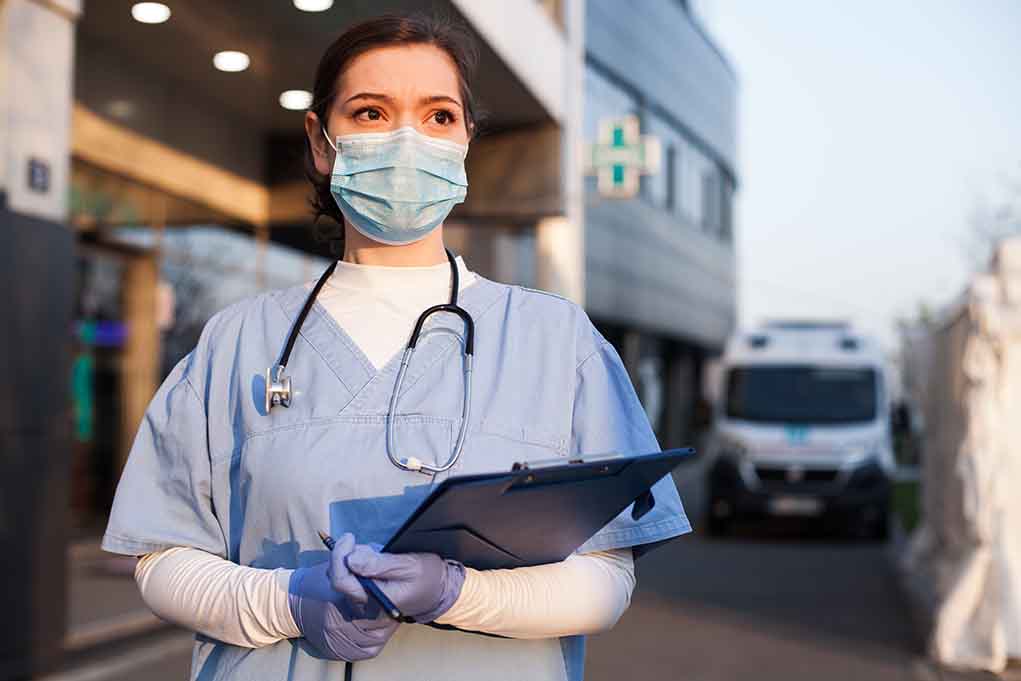 1714681855 featured image Doctor wearing mask and gloves, holding clipboard outside hospital.