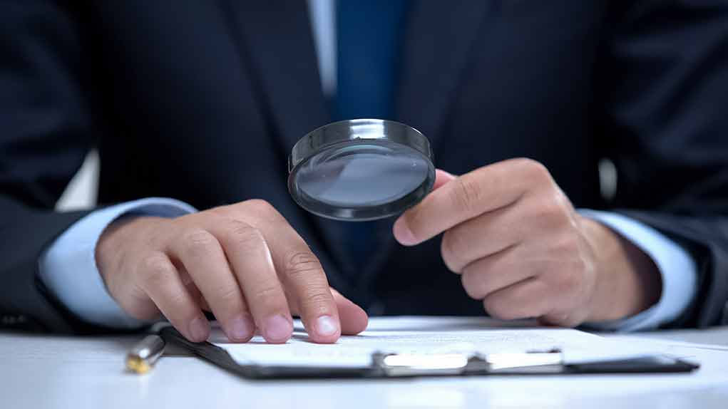 1539656501 featured image Man holding magnifying glass, examining document on clipboard.