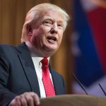 Exploring Trump’s Leadership Impact on Cabinet Autonomy and Efficiency Man in suit speaking at podium with flag background