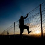 Silhouette of person climbing barbed wire fence at sunset