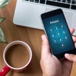 Typing privacy passcode on smartphone near laptop and coffee
