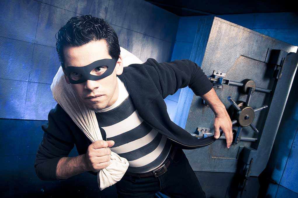 shutterstock_75825736.jpg A masked thief in a striped shirt holding a bag while emerging from a bank vault