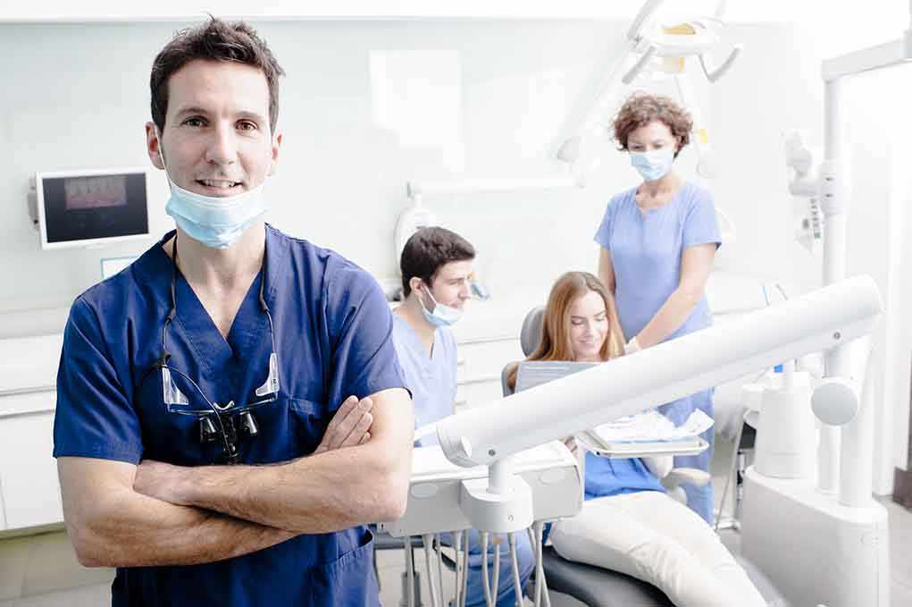 A dentist smiling in a dental office with a team assisting a patient