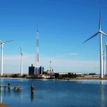 Wind turbines and a communication tower near a body of water under a clear blue sky