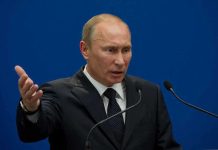 A man in a suit gesturing while speaking at a podium with microphones against a blue background