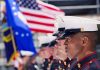 Marines in uniform standing in formation with flags in the background