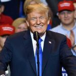 Trump’s Name Reappears In Epstein Flight Records Man in suit speaking at rally with red hats.