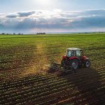 Farmer Crisis Forces Trump’s Hand Tractor plowing a vast green field at sunset.