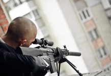 Person aiming a rifle from a building balcony.