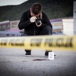Crime scene photographer behind police tape with evidence marker.