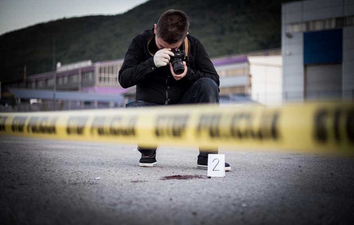 Crime scene photographer behind police tape with evidence marker.