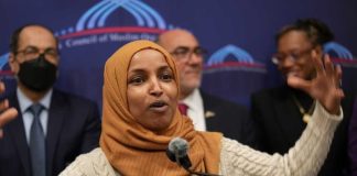 Ilhan Omar Firestorm Morphs Into Something Bigger A woman in a hijab passionately speaking at a podium with a diverse group in the background