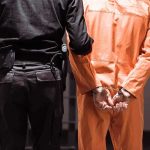 Abbott Refuses Minnesota’s ICE Demand: ‘I Don’t Trust Walz’ Prisoner in orange jumpsuit handcuffed with guard standing beside
