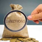 Trump Launches ‘Most Crushing’ Economic Attack on Iran hand holding magnifying glass over burlap money bag with coins
