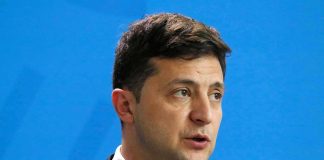 Volodymyr Zelensky speaking at a podium against a blue backdrop