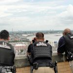 9 Officers Plunge as Deck Collapses During Raid Three U.S. Marshals in tactical vests overlooking a city and river