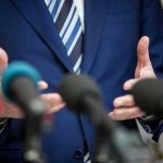 Speaker in blue suit gesturing behind microphones