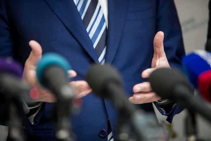 shutterstock_1670760397 Speaker in blue suit gesturing behind microphones