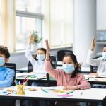Families Could Pay the Price for Governor’s Decision Schoolchildren wearing masks raise hands in a classroom
