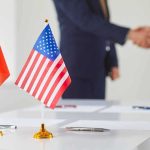 Chinese and American flags on a table with a handshake in the background