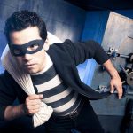 A masked thief in a striped shirt holding a bag while emerging from a bank vault