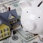 A model house next to a white piggy bank on a bed of dollar bills